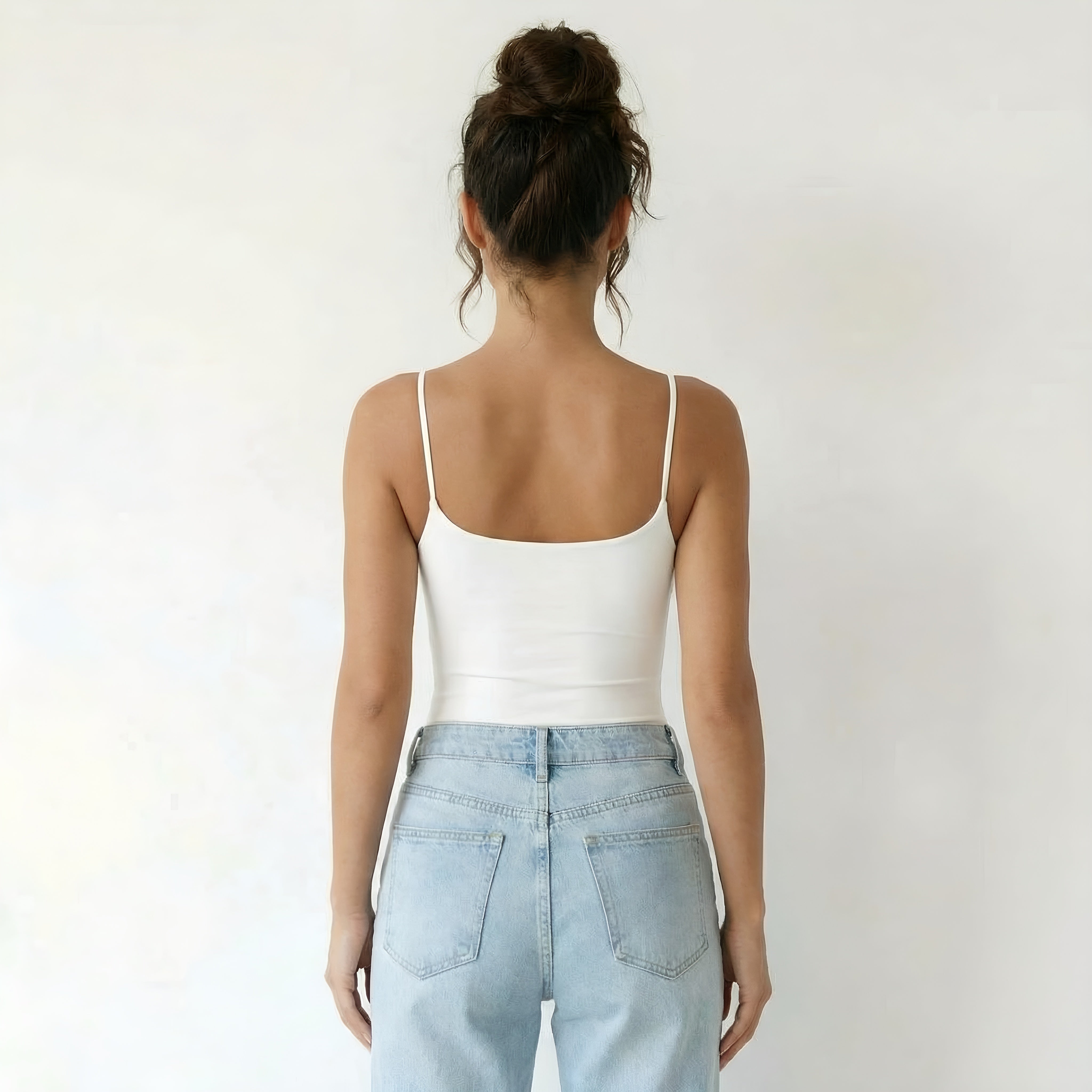Woman wearing a white tank top and light blue jeans against a plain. White shapewear top lyvia background
