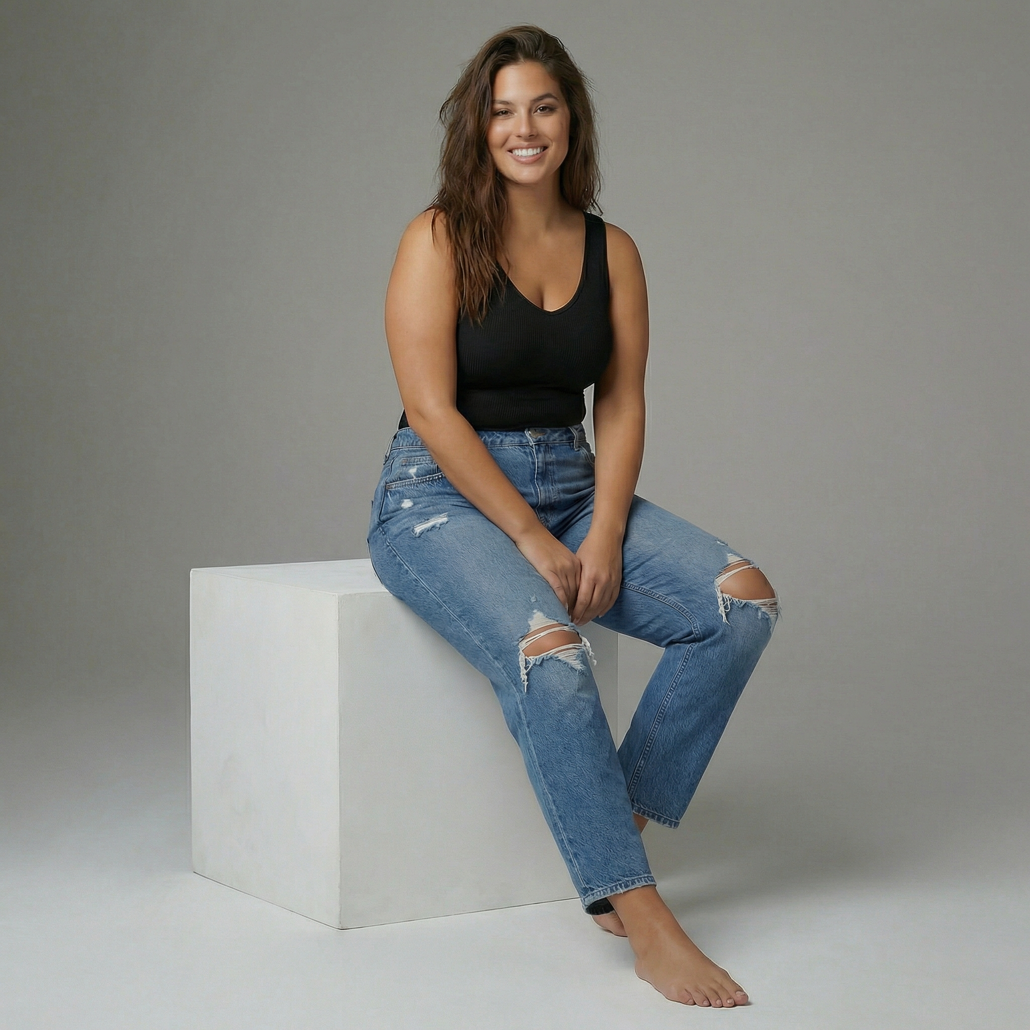 Woman sitting on a white cube wearing a black tank top and ripped blue jeans against a gray background black shapewear womans lyvia